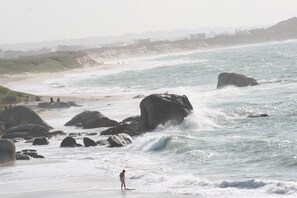 A beach with large rocks and waves, a person standing on a surfboard, and a distant shoreline with buildings.