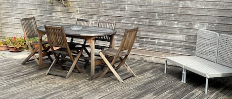 Um deck de madeira com uma mesa redonda e cadeiras, um banco e um muro baixo com plantas.