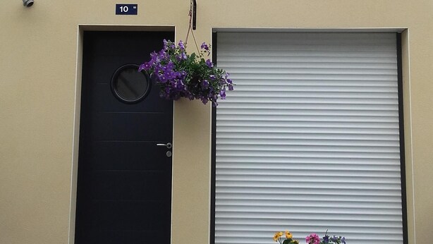 A building with a black door, a number '10' sign, a window with hanging flowers, and a closed roller shutter. - duplex apartment bis (Vannes)