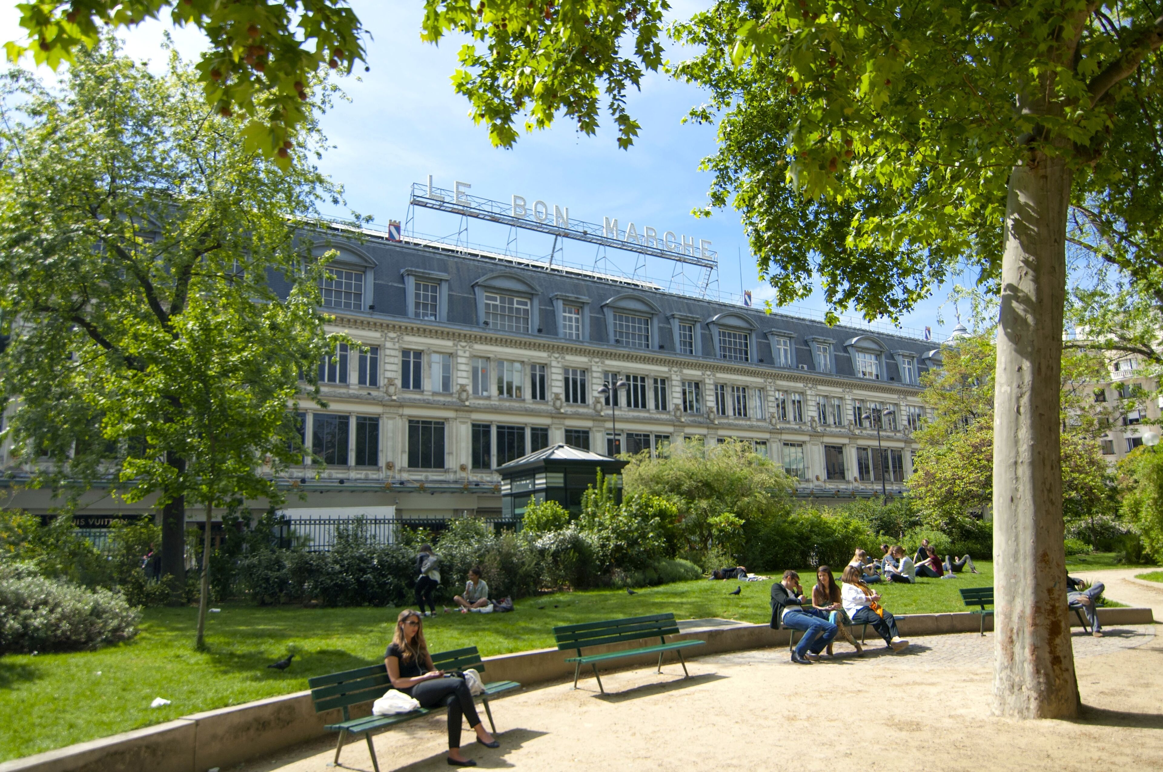 Ein Park mit Bänken und sitzenden Personen. Im Hintergrund ein großes Gebäude mit der Aufschrift 'LE BON MARCHE'.
