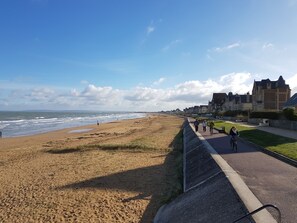 A coastal walkway with a sandy beach, a paved path, and buildings in the background. - Villa Le Petit Bois - 2 mns walk Sword beach -Large enclosed garden - Car park (Hermanville-Sur-Mer)