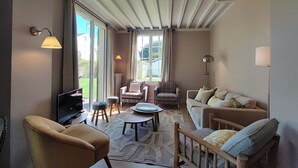 A spacious living room with a large window, a sofa, a chair, a coffee table, and a TV. - Villa Le Petit Bois - 2 mns walk Sword beach -Large enclosed garden - Car park (Hermanville-Sur-Mer)
