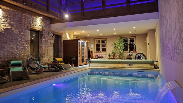 Una piscina interior con cubierta de madera y sillas de descanso, rodeada de muros de piedra y techo de madera.