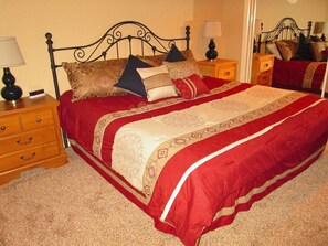 Een net opgemaakt bed met een rood-beige sprei, twee nachtkastjes met lampen en een houten dressoir.