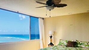 Une chambre avec une grande fenêtre donnant sur l’océan, un lit recouvert d’une courtepointe à motifs tropicaux, un ventilateur de plafond et une table de chevet avec une lampe.