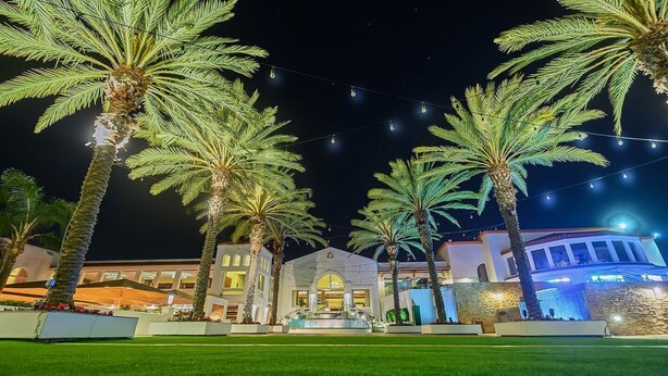 Un hotel con una entrada bien iluminada, palmeras y un área con césped.
