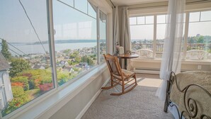 A room with a large window offering a view of a body of water and a cityscape. There is a rocking chair, a small table with a lamp, and a bed with a canopy.