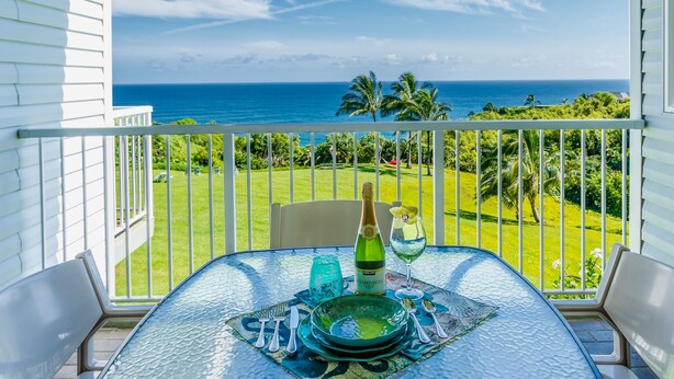 A glass-top table with a bottle of champagne, a glass, and cutlery on a patterned table runner, overlooking a lush green lawn and the ocean. - Very Private location. Amazing View. The Cliffs 6302, 1 Vehicle Parking Included (Princeville)