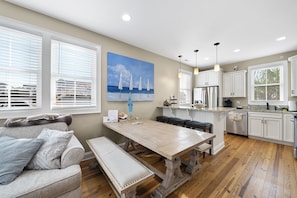 A spacious kitchen with white cabinets, a wooden dining table, and a bench. There is a large window with blinds, a painting of a beach scene, and a view of houses outside.