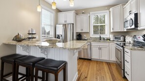 Una cocina con armarios blancos, un refrigerador de acero inoxidable, un microondas y una encimera de granito. Hay tres taburetes negros y luces colgantes sobre la isla.