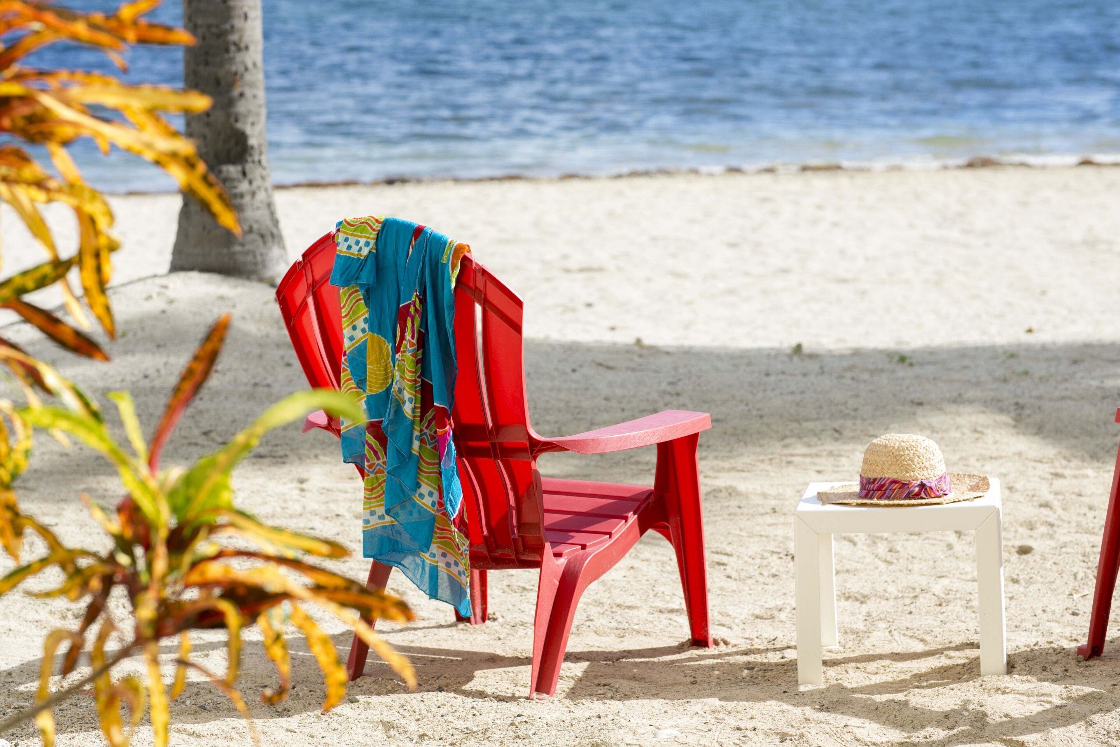 Une chaise de plage rouge avec un tissu aux couleurs vives, une petite table blanche surmontée d’un chapeau de paille, et une plage de sable avec un palmier et l’océan en arrière-plan.