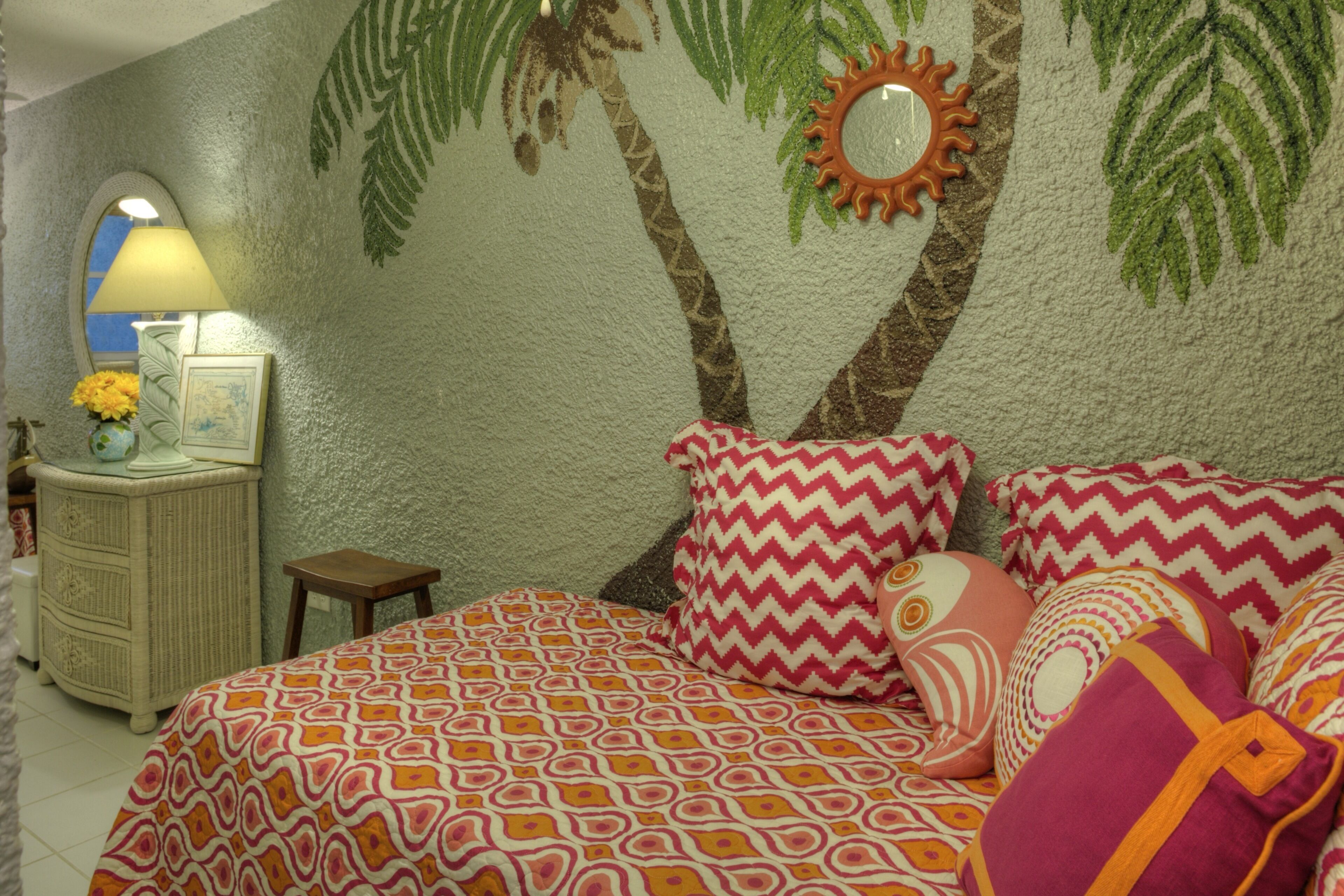 Un dormitorio con cama, almohadas, una mesita de noche, una lámpara y un mural en la pared con palmeras.