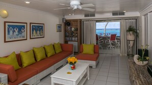 A living room with a white ceiling fan, a large sofa with green cushions, a white coffee table with a vase of yellow flowers, and a view of the ocean through a glass door.