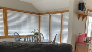 A living room with a black leather sofa, a blue pillow with text, a dining table with chairs, and large windows with blinds. - Muskoka Cottage Living  (Huntsville)