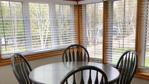 A corner dining area with a round table and six chairs, large windows with blinds, and wooden flooring. - Muskoka Cottage Living  (Huntsville)