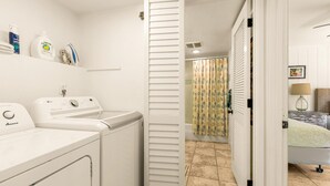 A laundry area with a front-loading washer and dryer, a shelf with cleaning products, and a bathroom with a shower curtain and towel rack.