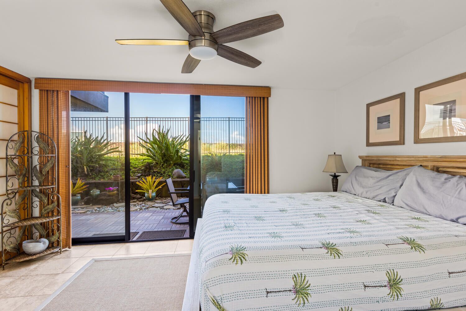 A bedroom with a bed, a ceiling fan, a nightstand, a lamp, and a view of a garden through a sliding glass door.