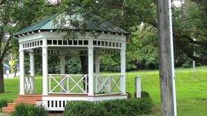 Een witte pergola met een groen dak, omringd door weelderige begroeiing en een houten omheining.