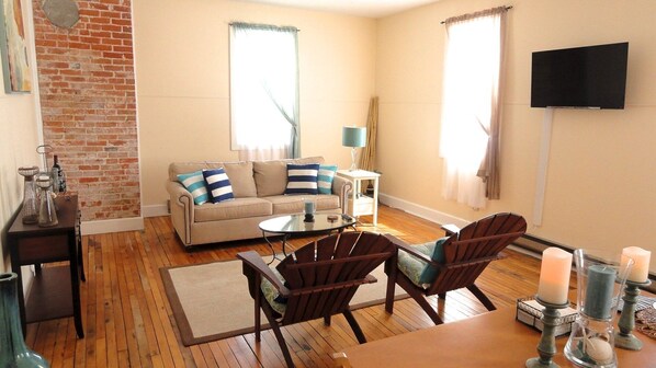 A cozy living room with a brick wall, a beige sofa, a coffee table, two wooden chairs, a small side table, and a TV mounted on the wall. - Marine on St Croix Retreat (Marine On Saint Croi)