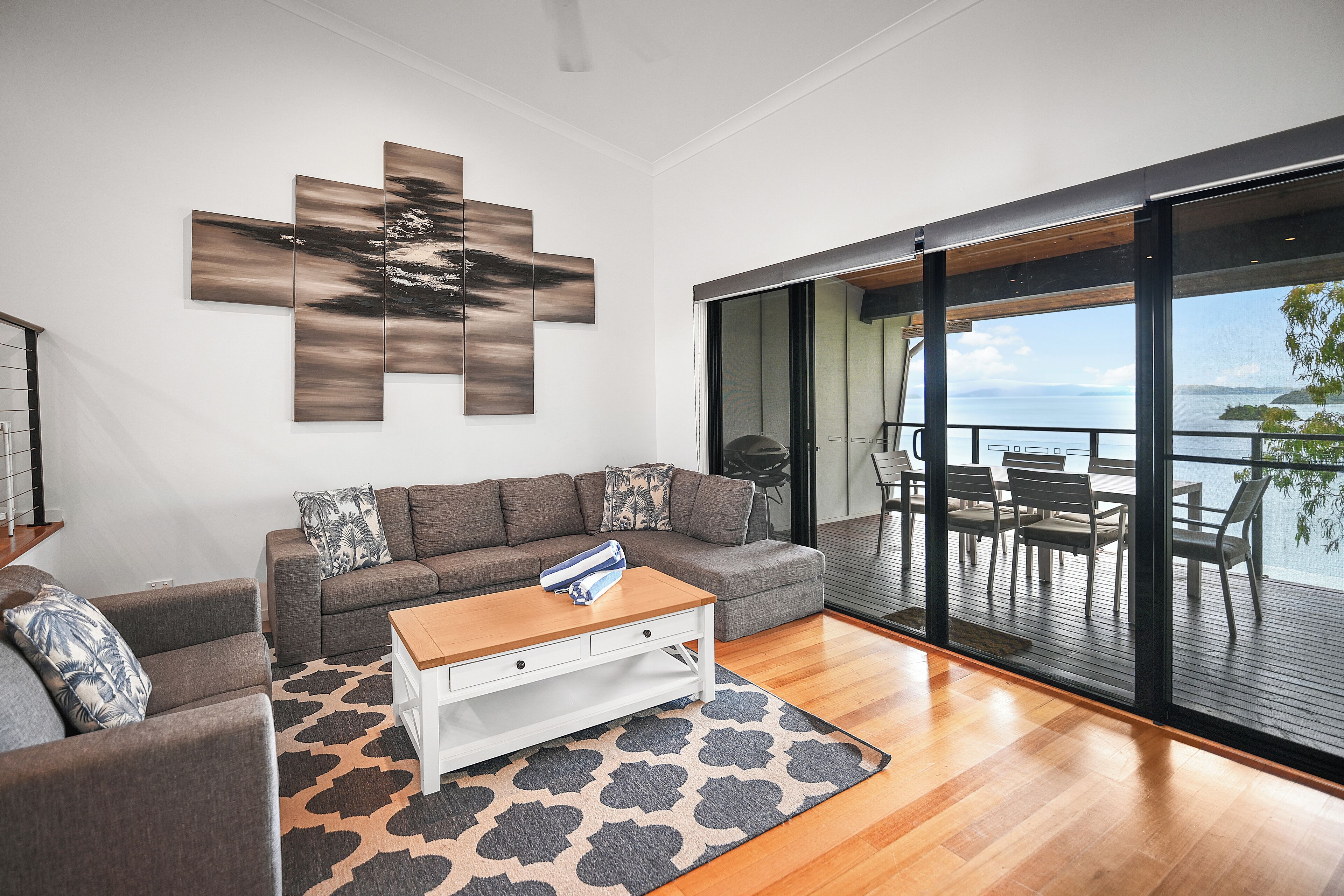 A modern living room with a sofa, coffee table, and a large window overlooking a deck with outdoor furniture.