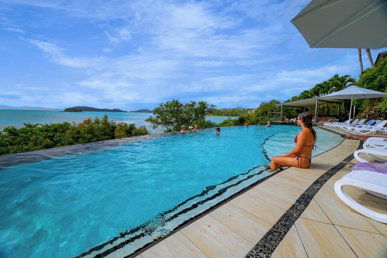 Zona junto a la piscina con vista al mar, sillas de descanso y una persona sentada a la orilla.