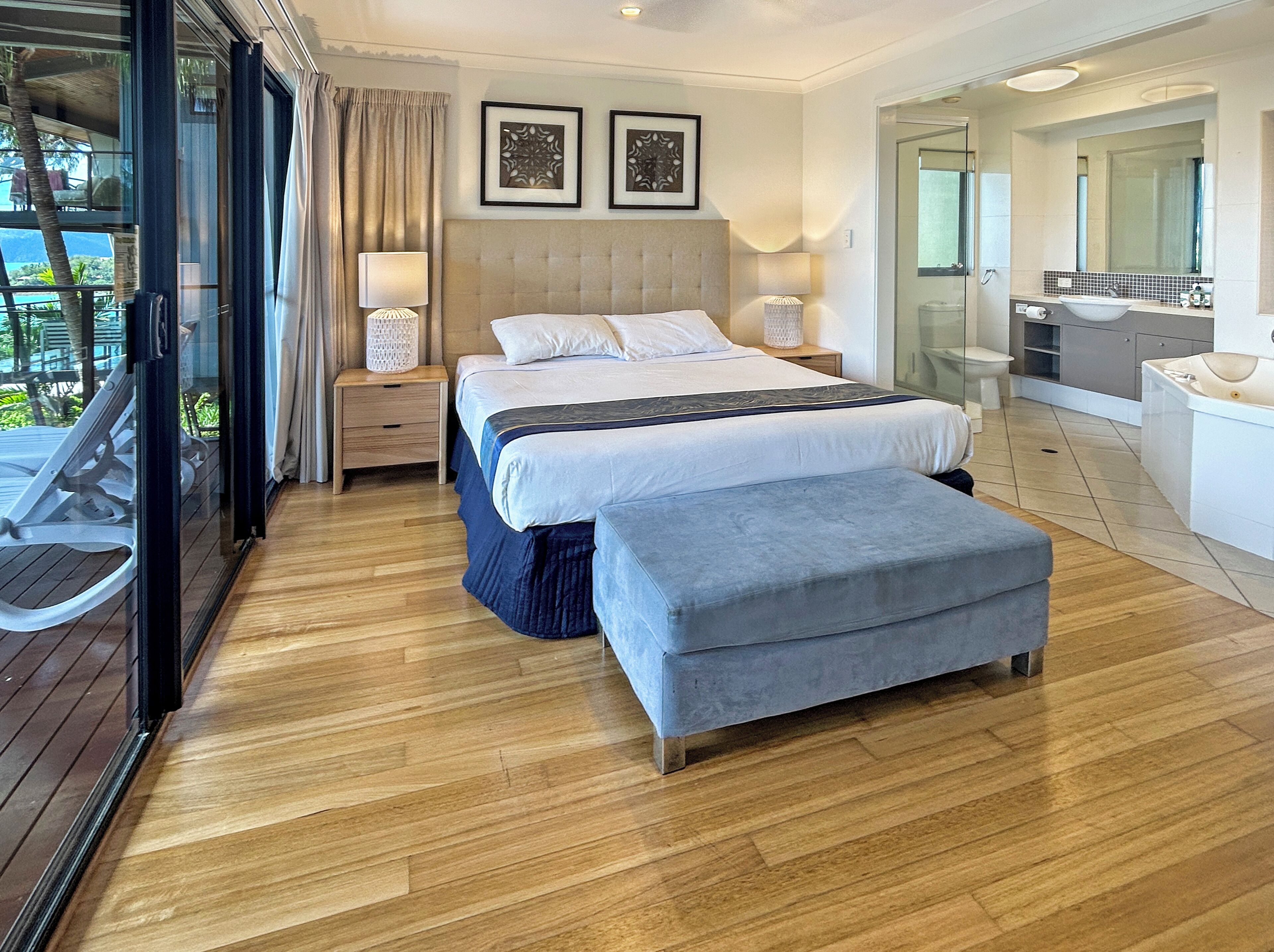 A modern hotel room with a large bed, a sitting bench, a balcony with a view, a bathroom with a large sink and mirror, and two framed pictures on the wall.