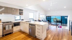 A modern kitchen with a central island, built-in oven, microwave, and dishwasher. It features wooden flooring, white cabinetry, and a large window offering a view of the sea. - Shoreline Apartments (Whitsundays)