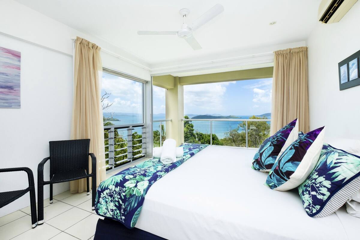 A bedroom with a large bed, a ceiling fan, and a view of the sea.