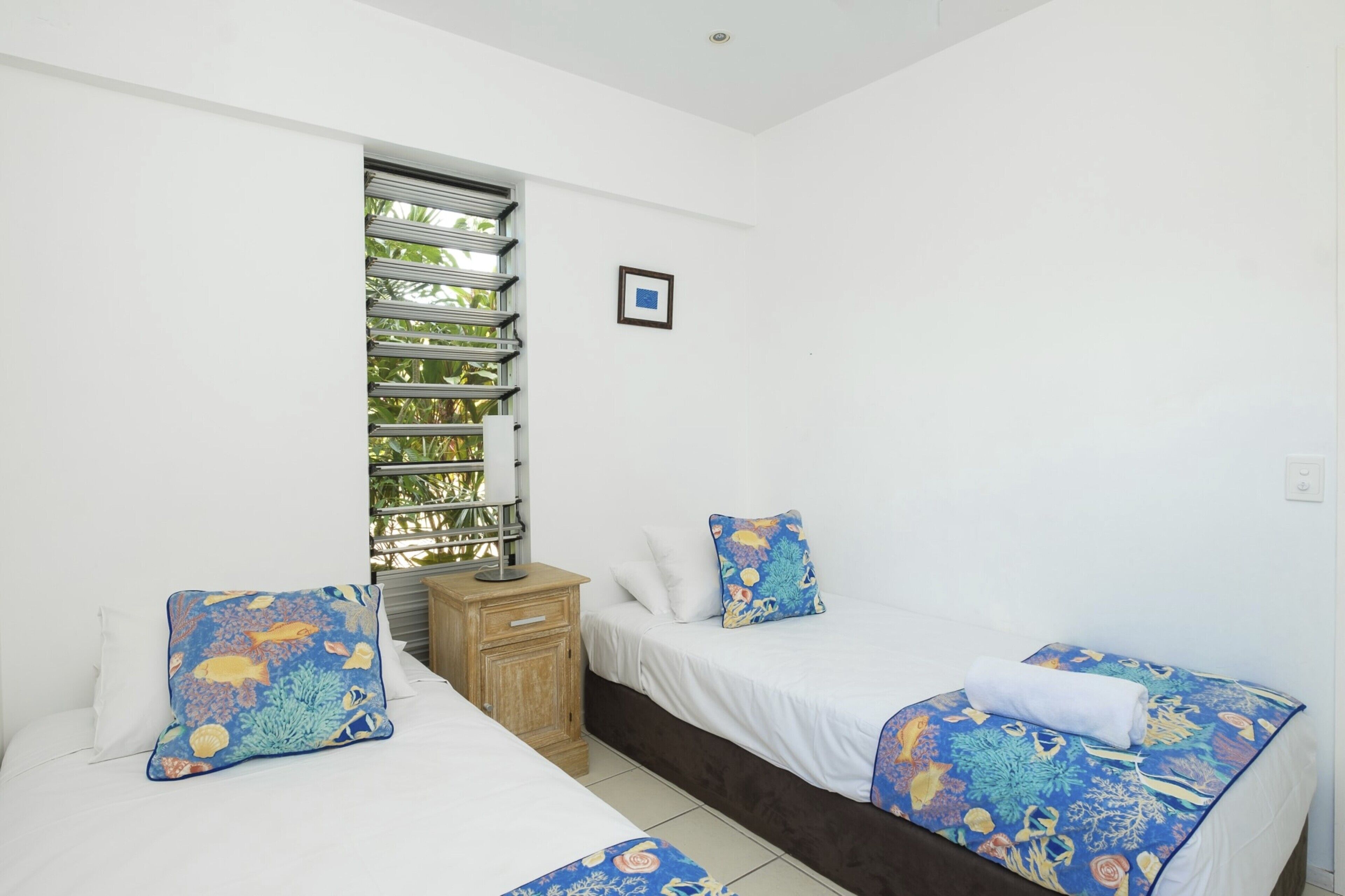 A room with two beds, a window with blinds, a small wooden nightstand, and a framed picture on the wall.