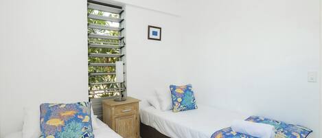 A room with two beds, a window with blinds, a small wooden nightstand, and a framed picture on the wall.