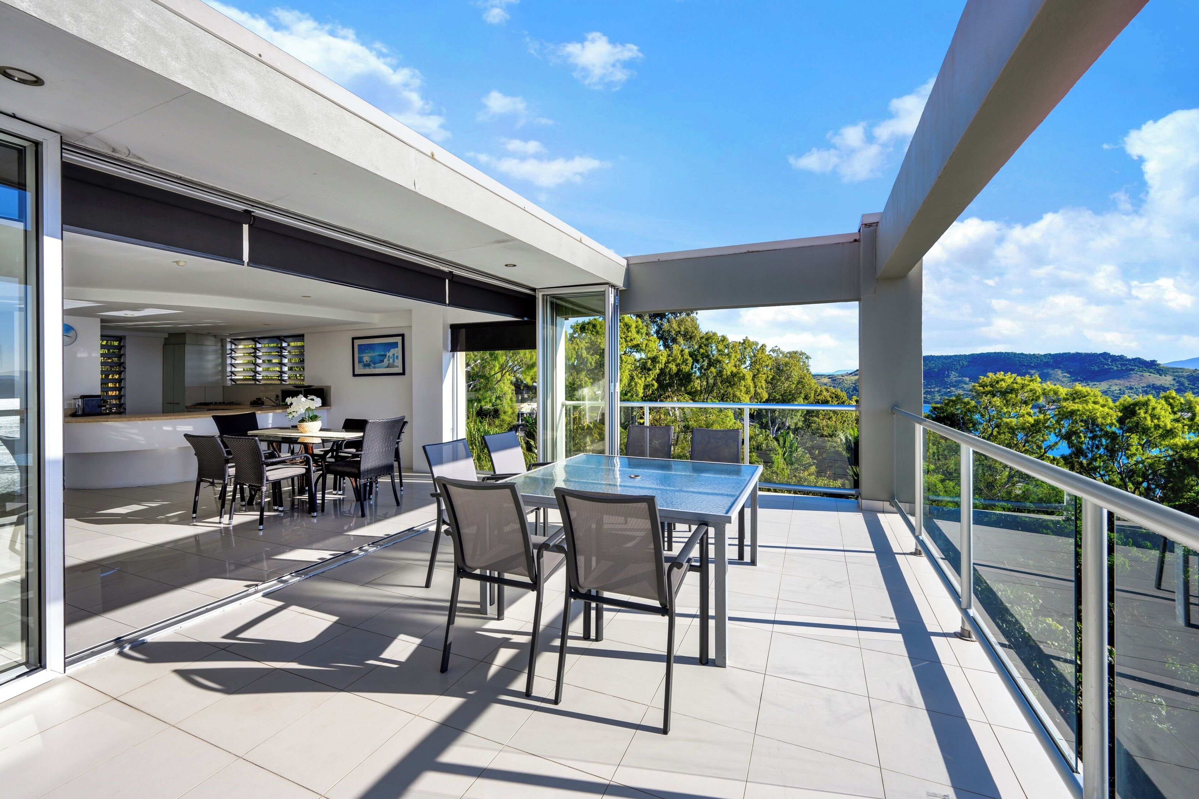 A modern outdoor dining area with a glass table and chairs, a kitchen area with a dining table and chairs, and a view of the surrounding landscape.
