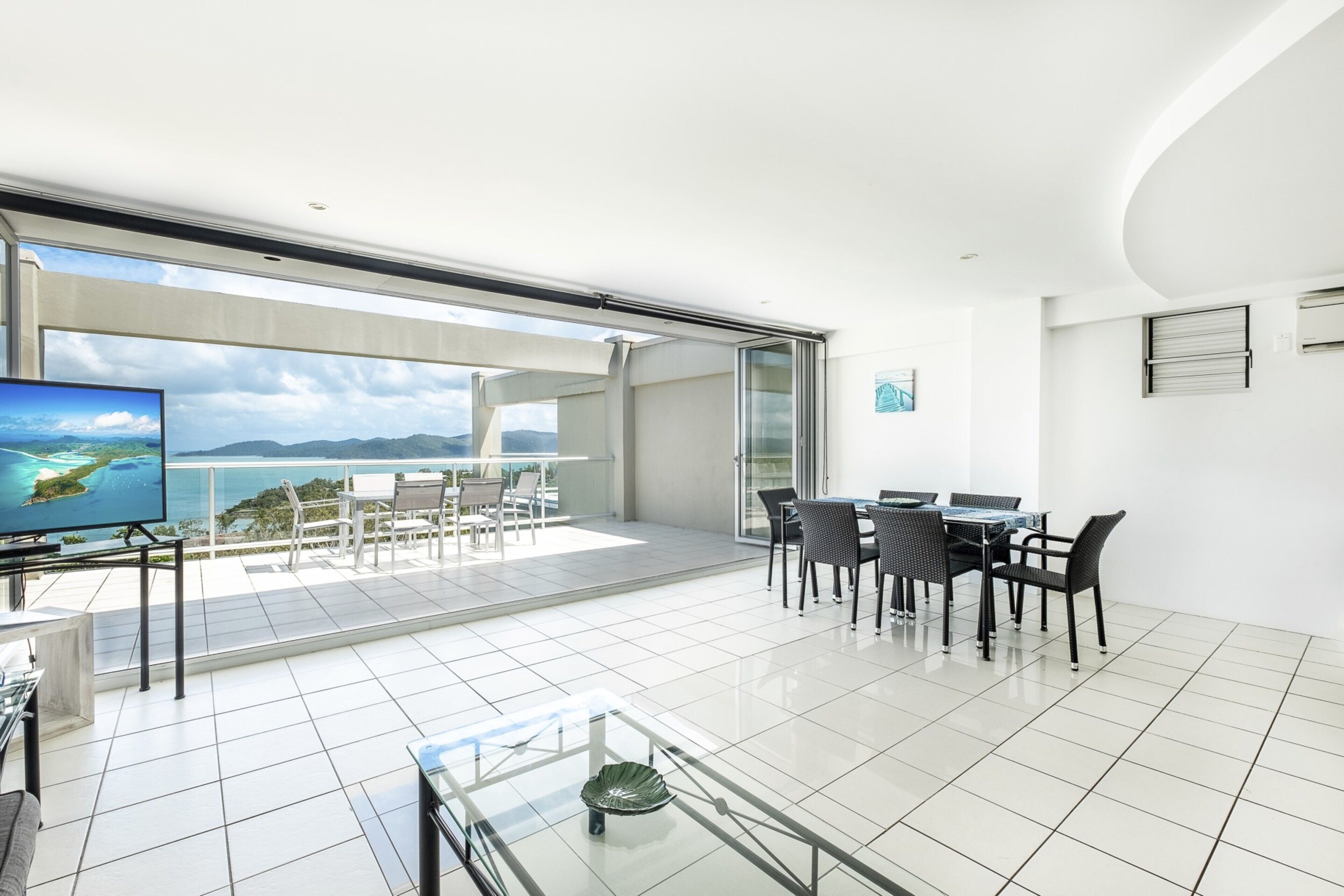 A spacious living area with a large glass door leading to a balcony, a dining table with chairs, and a flat-screen TV.