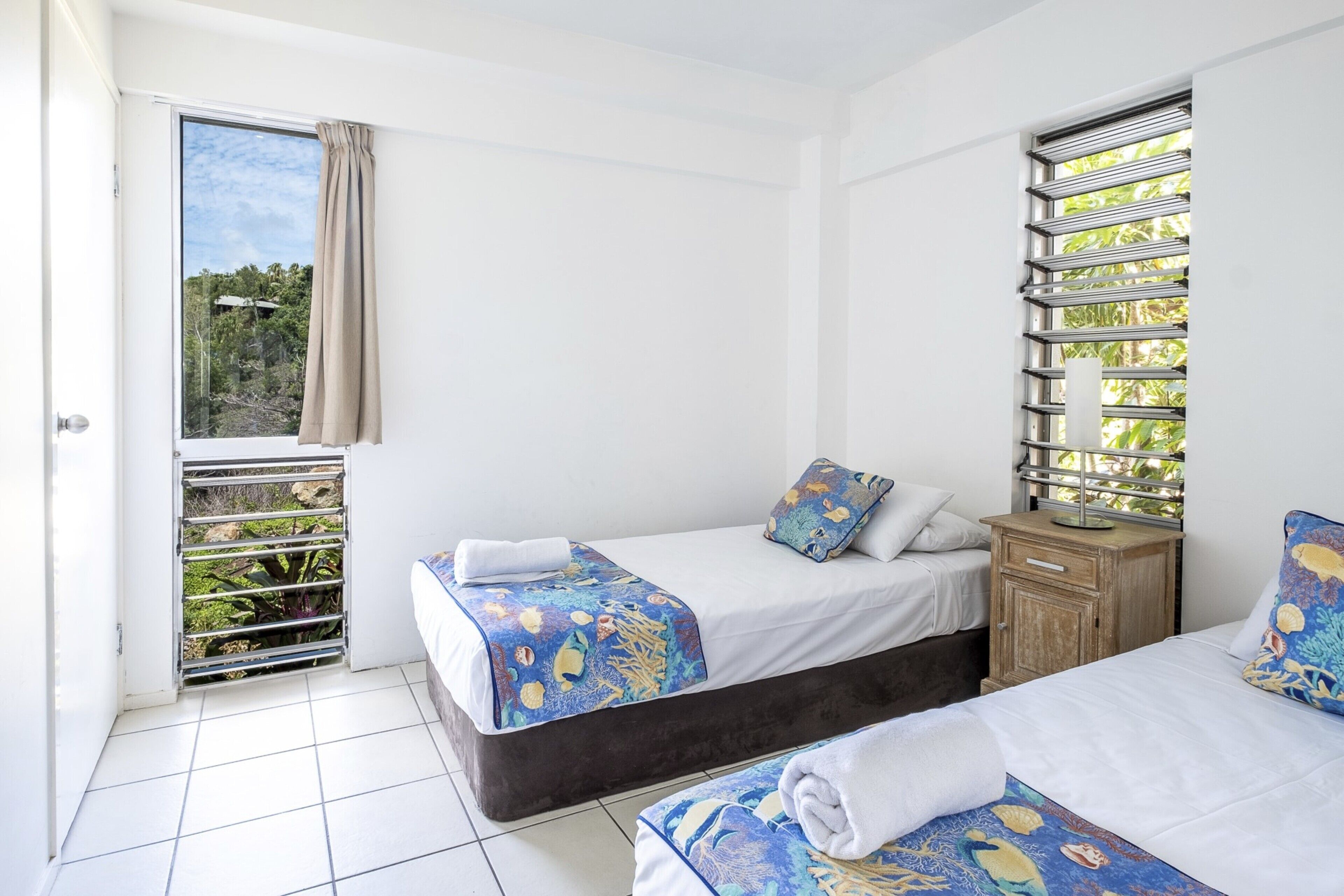 A room with two beds, a window with a metal grill, a wooden nightstand, and a balcony door.
