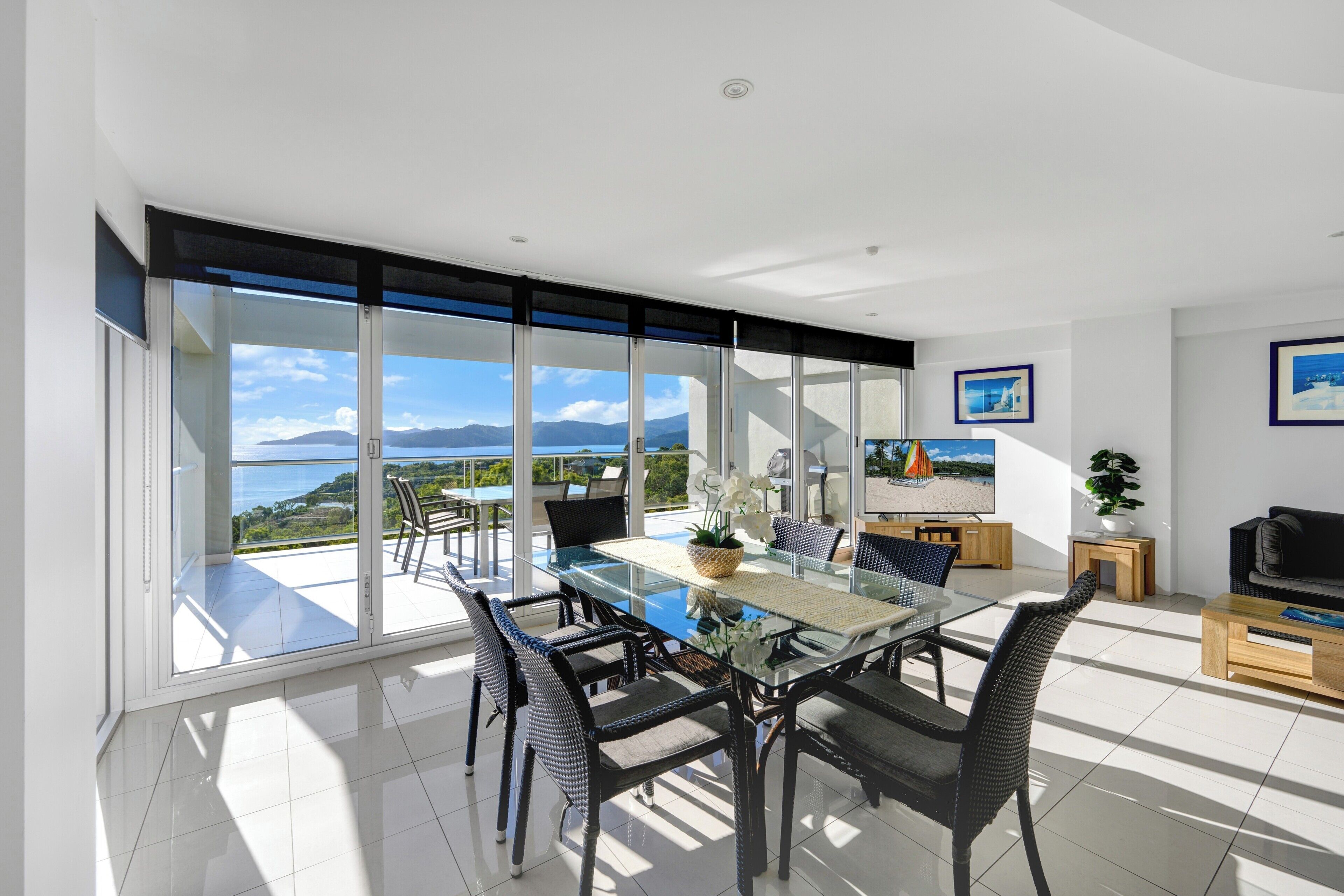 A modern living room with a glass dining table, black wicker chairs, a sofa, a TV, and a view of the mountains.