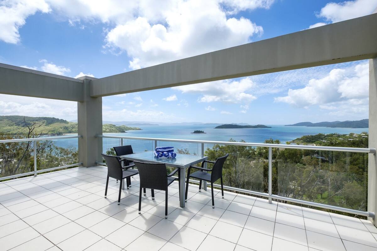A balcony with a table and chairs, offering a view of a coastal landscape with hills and a body of water.
