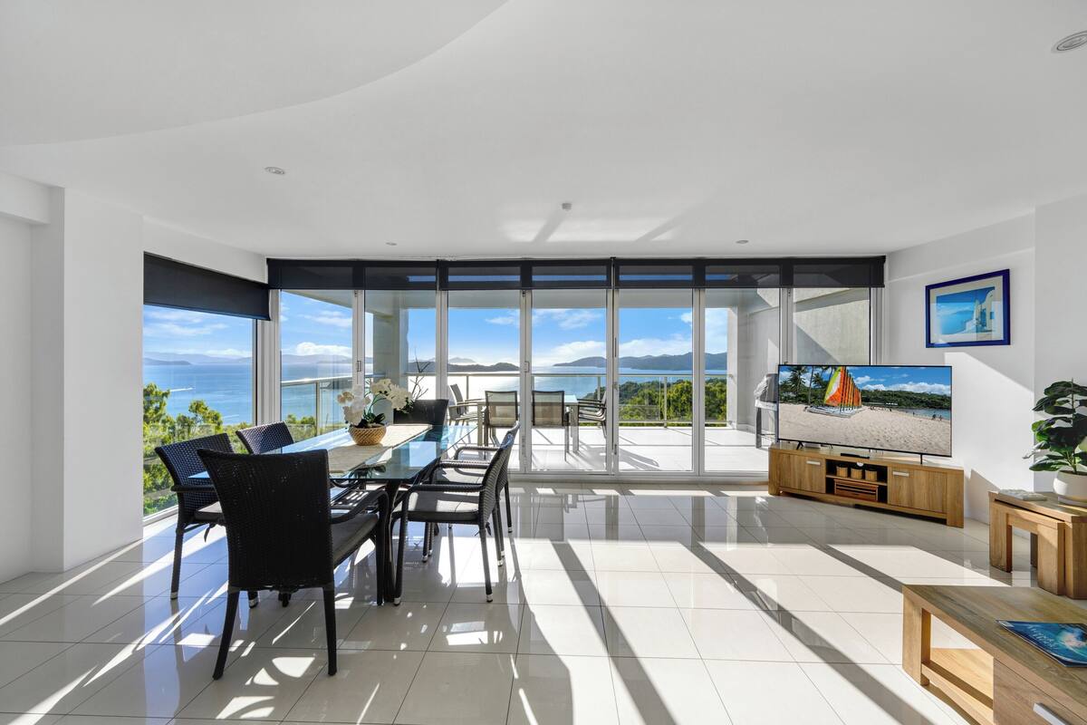 A modern living room with a large TV, dining area, and a view of the ocean.