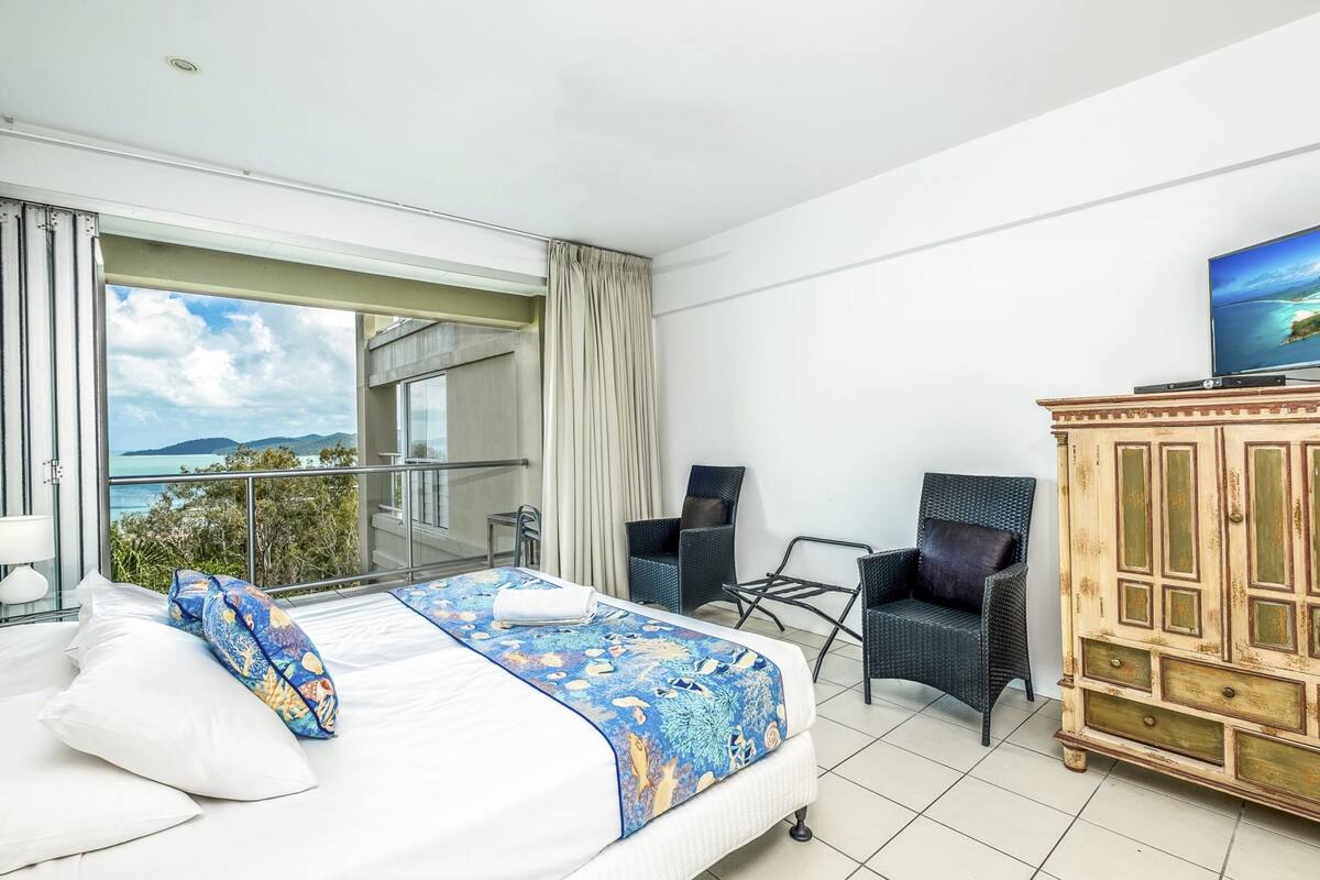 A hotel room with a bed, two chairs, a wooden cabinet, and a TV.