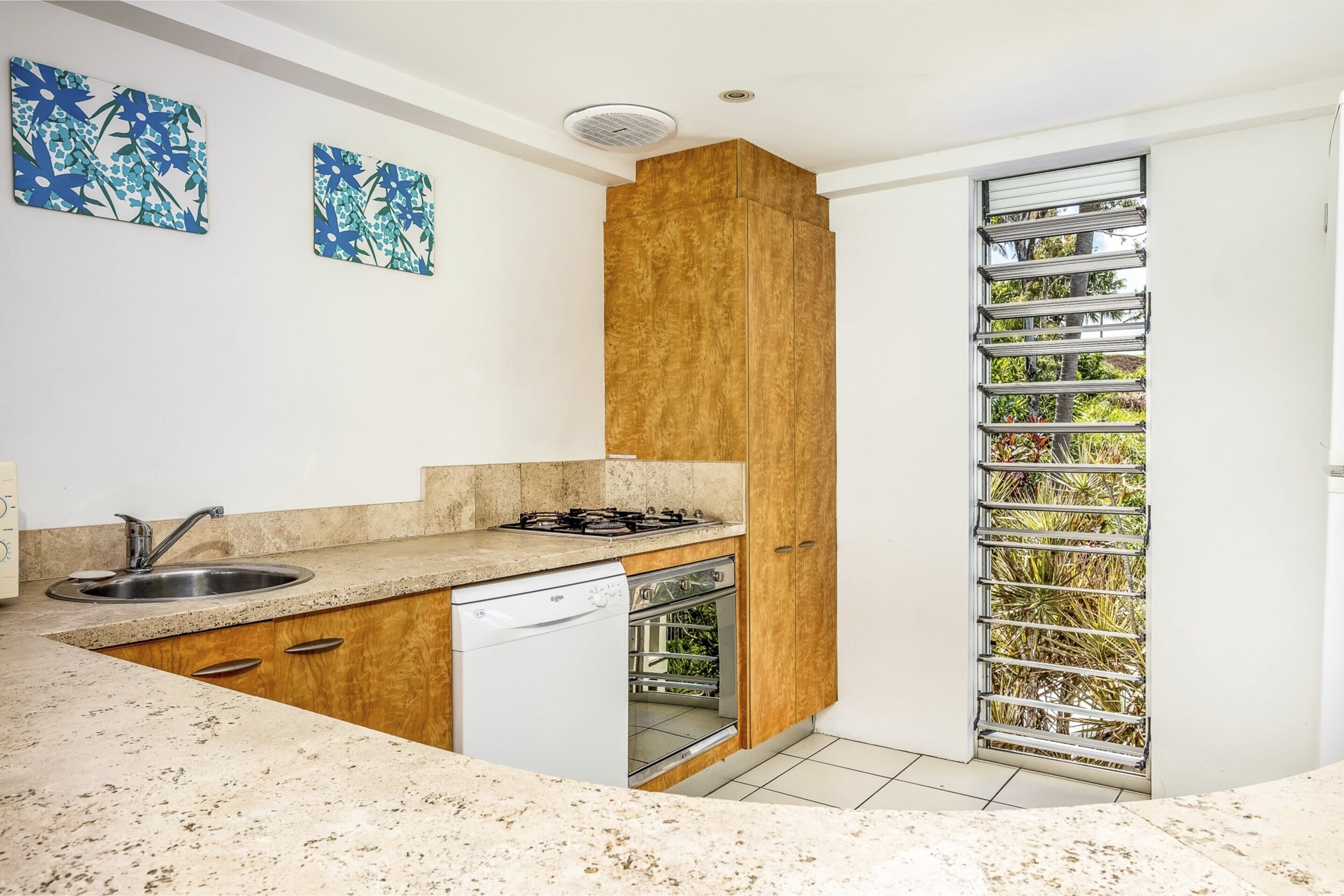 A modern kitchen with a built-in dishwasher, a stove, and a sink. There are two wall-mounted artworks and a window with a metal grill.
