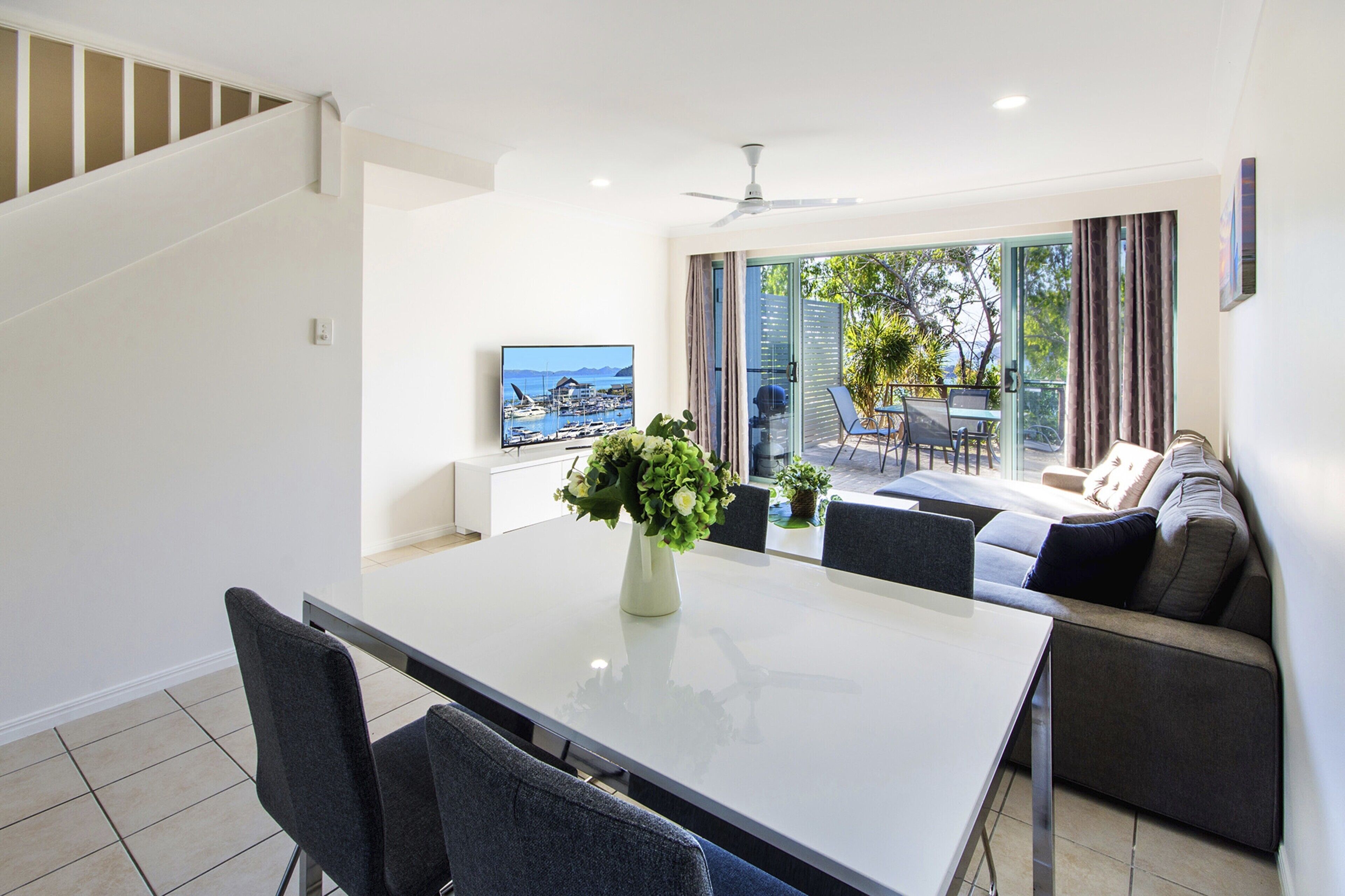 Un comedor moderno con una mesa blanca, sillas grises y un florero. Un televisor de pantalla plana está montado en la pared, y se ve un sofá al fondo.