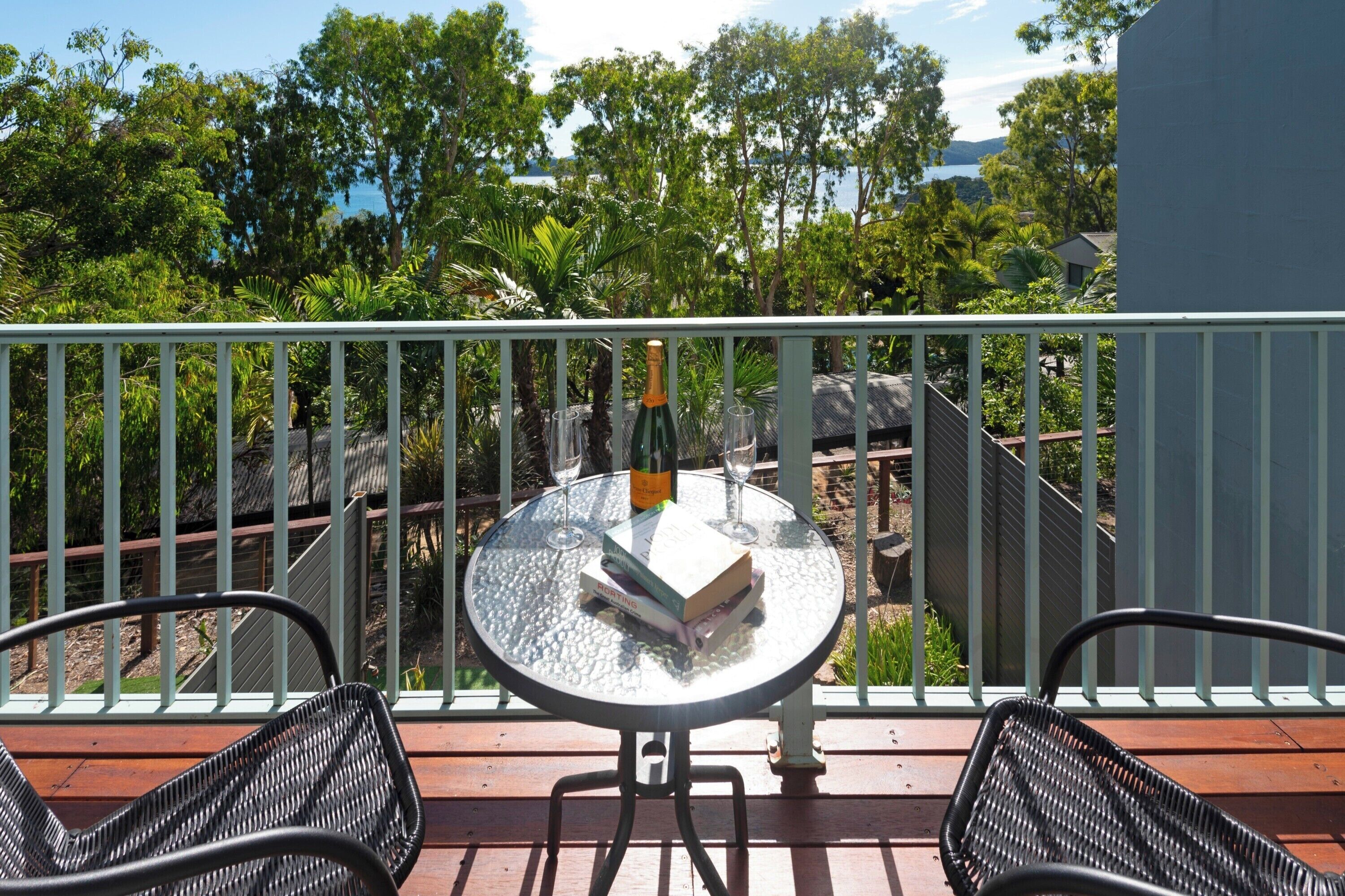 A balcony with a round glass table, two wicker chairs, a bottle of champagne, and two wine glasses.