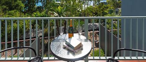 A balcony with a round glass table, two wicker chairs, a bottle of champagne, and two wine glasses.
