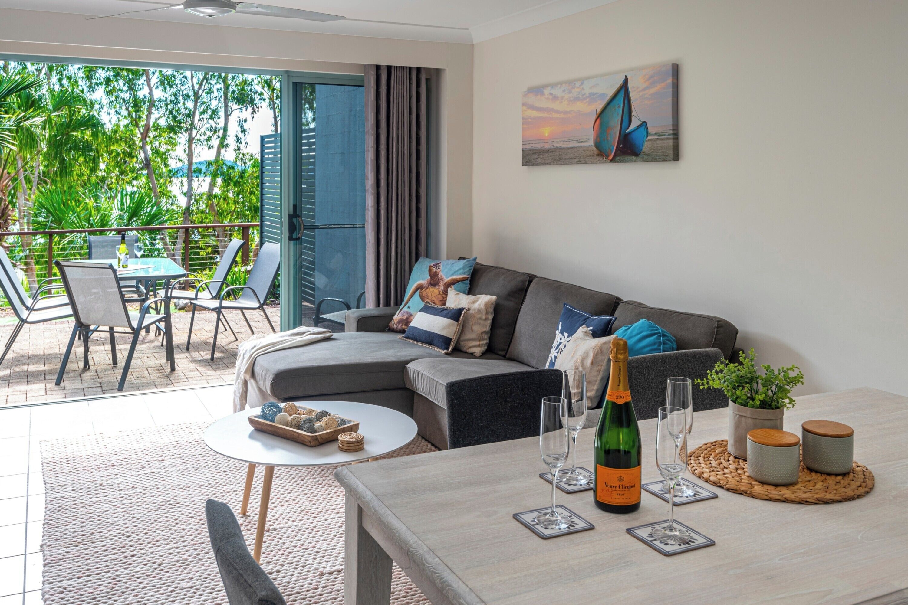 A modern living room with a grey sofa, a dining table with a bottle of champagne, and a view of the outdoors through large windows.