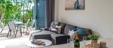 A modern living room with a grey sofa, a dining table with a bottle of champagne, and a view of the outdoors through large windows.