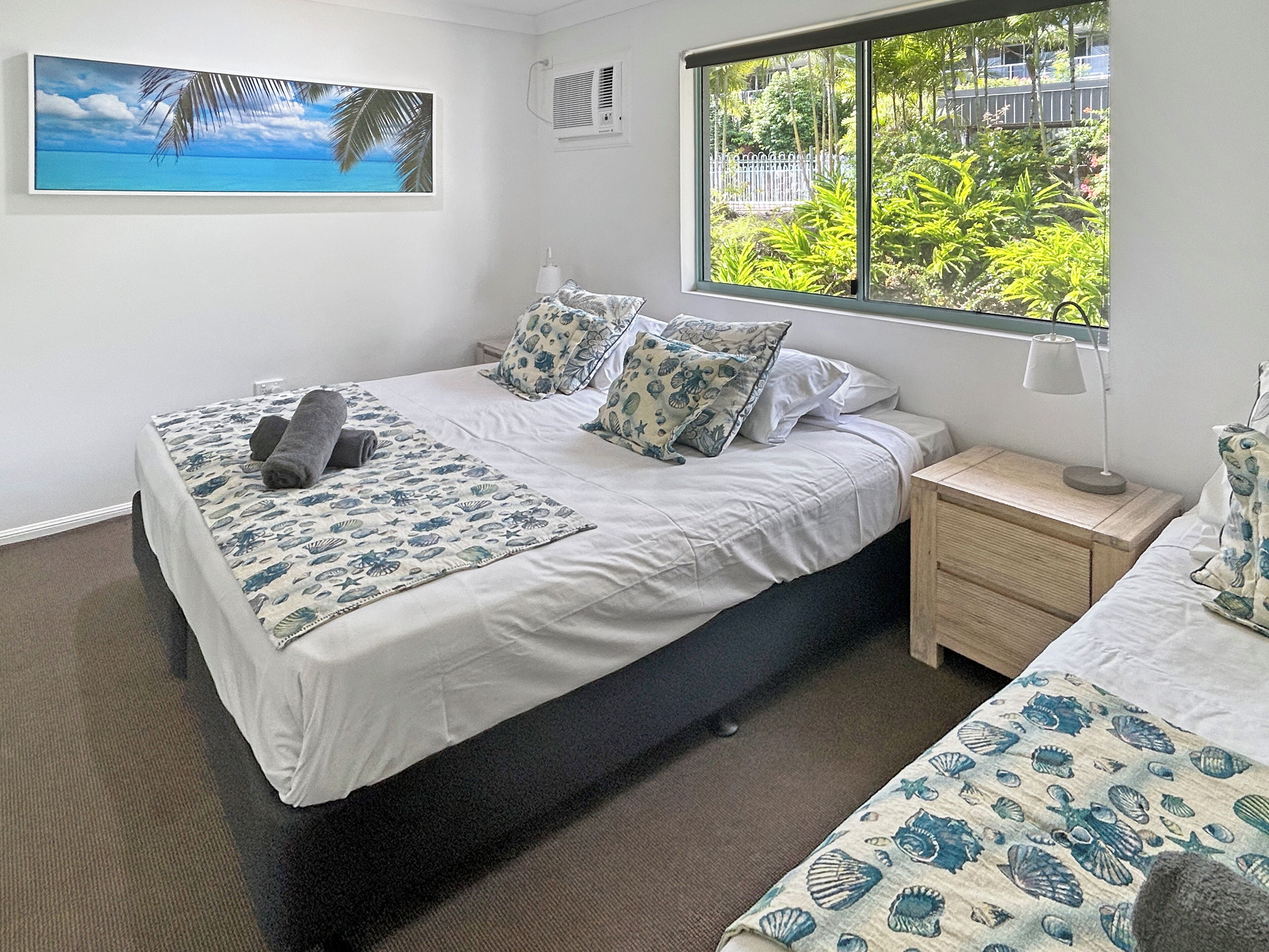 A bedroom with a bed, a nightstand, a window with a view of greenery, and a wall-mounted air conditioning unit.