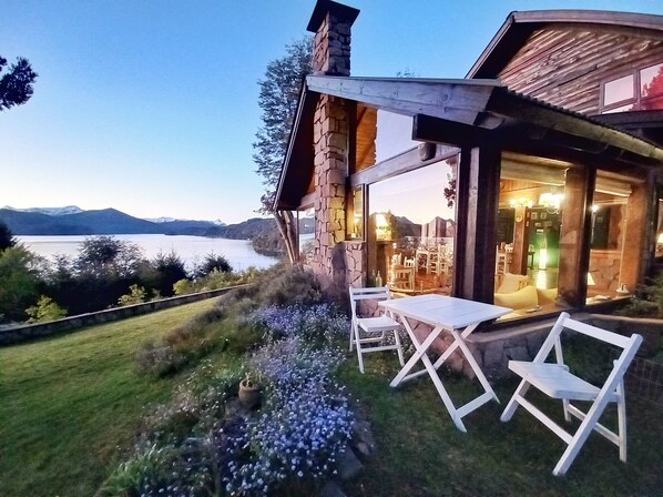 A stone building with a glass door and windows, a patio with a table and chairs, and a view of a lake and mountains in the background.
