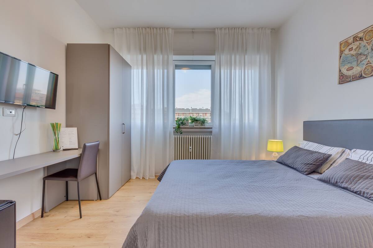 A modern hotel room with a large bed, a desk with a chair, a television, and a window with curtains.