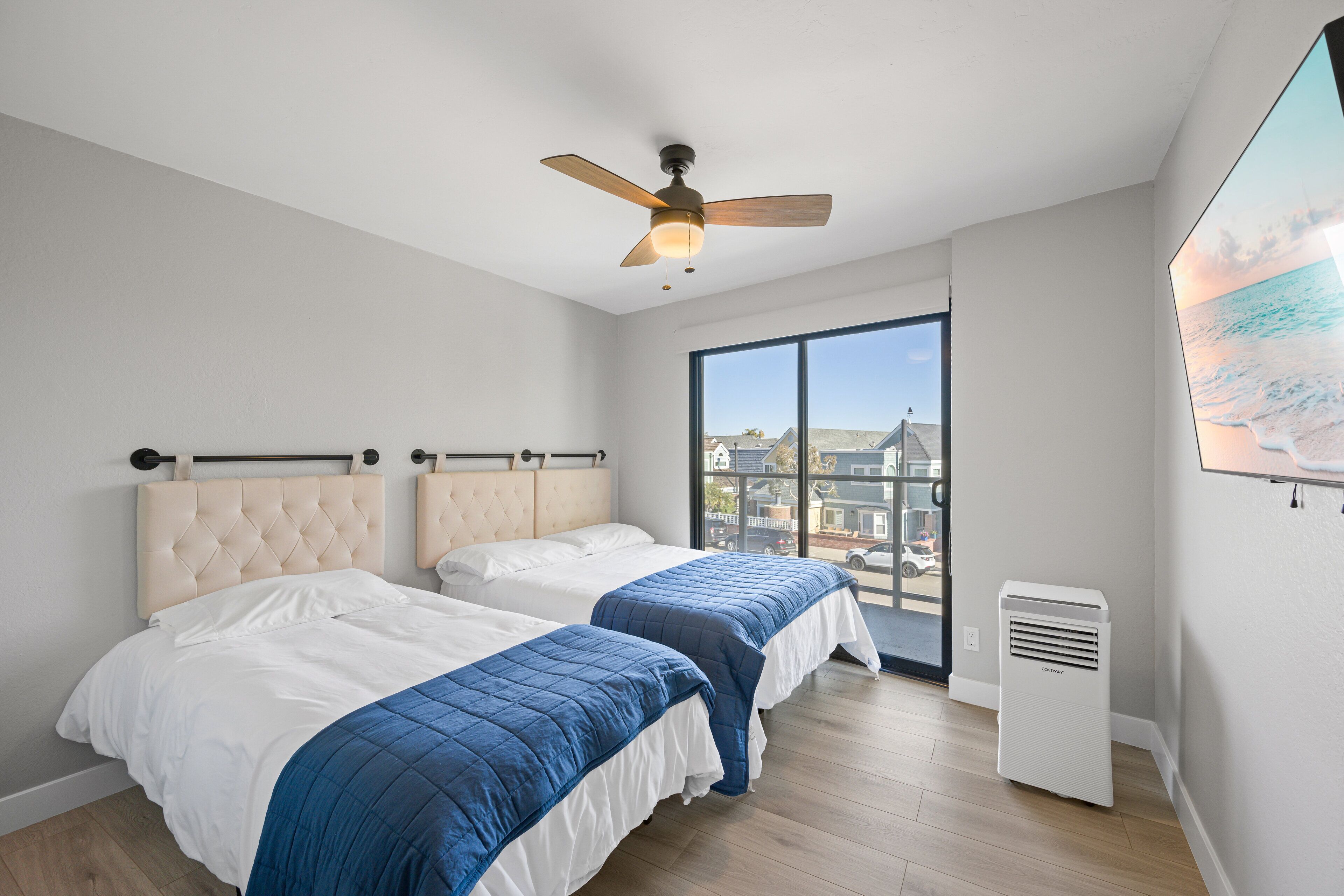 A bedroom with two beds, a ceiling fan, a large window with a view of buildings, and a wall-mounted air conditioner.