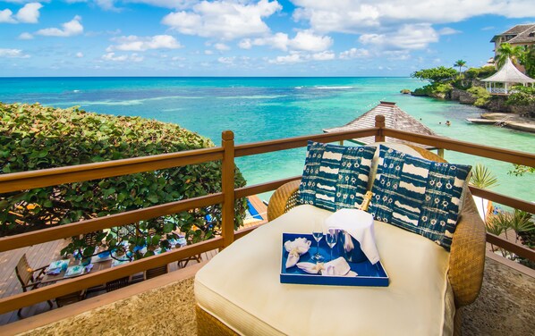 A balcony with a view of the ocean, a bed with a pillow, and a table set with a glass of water and a napkin.