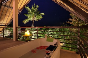 A private balcony with a bathtub, a candle, and a view of a palm tree at night.