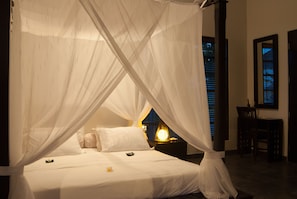 A bed with white linens and a canopy, a bedside table with a lamp, and a chair in a room with a dark floor and a window with blinds.
